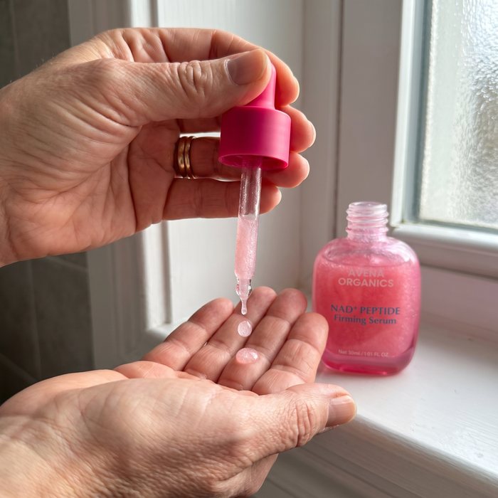 Drops of Avena NAD+ Peptide Serum in a woman’s palm, the open bottle on the windowsill