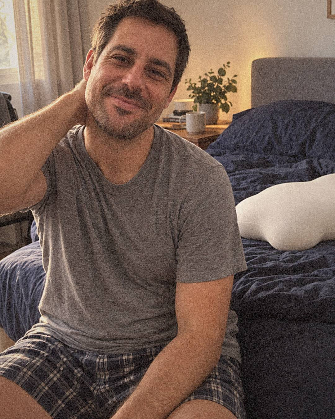 Marcus on the edge of his bed in the morning, stretching his neck and smiling, his Cloud Pillow behind him